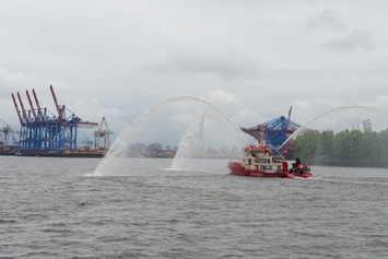 atlantis dx - 25 Jahre Firmenjubiläum Foto: Ein Löschboot spritzt Wasser auf einem Gewässer, im Hintergrund Hafenanlagen