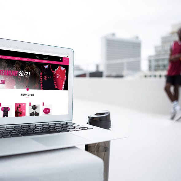 atlantis dx - EVENTIM SPORTS: Online-Shop-Plattform für Merchandising mit Magento 2 Foto: Outdooraufnahme, Laptop mit geöffnetem Shop von EVENTIM SPORTS, im Hintergrund ein Sportplatz ein Basketballspieler, ein weißes Hochhaus